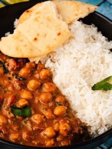 A black bowl with white rice, chickpea curry, and a piece of naan bread, garnished with cilantro, on a blue-tiled surface.