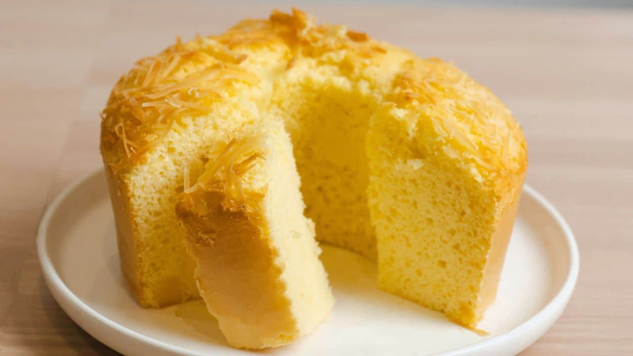 A round, fluffy sponge cake topped with grated cheese sits on a white plate with a large slice cut out.
