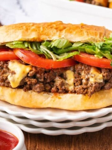 A sandwich with ground beef, melted cheese, tomato slices, and shredded lettuce on a bun, served on a plate with side dishes of mustard, ketchup, and a bowl of lettuce.