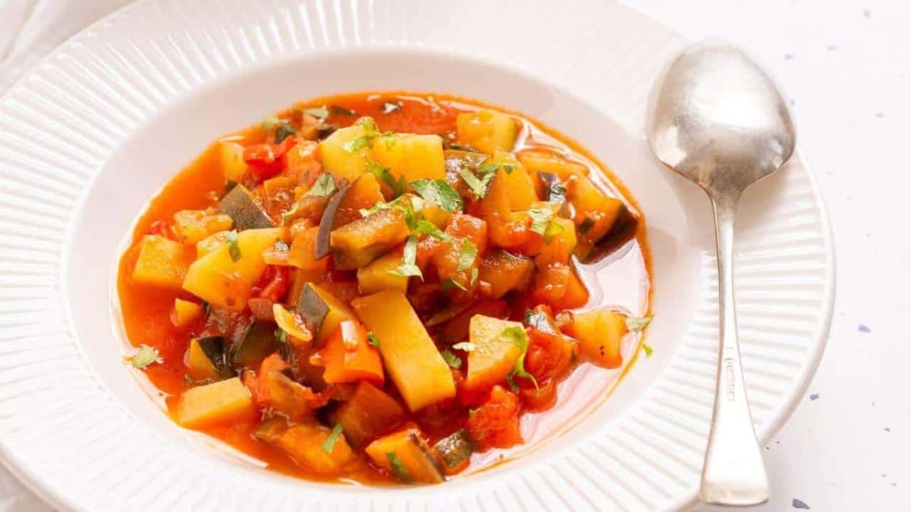 A pot of Italian vegetable stew with a spoon, garnished with chopped green herbs, on a white tablecloth with a small bowl of parsley on the side.