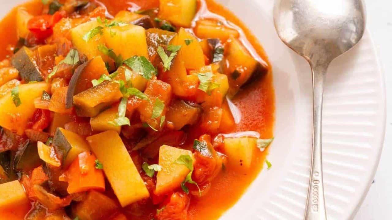 A pot of Italian vegetable stew with a spoon, garnished with chopped green herbs, on a white tablecloth with a small bowl of parsley on the side.