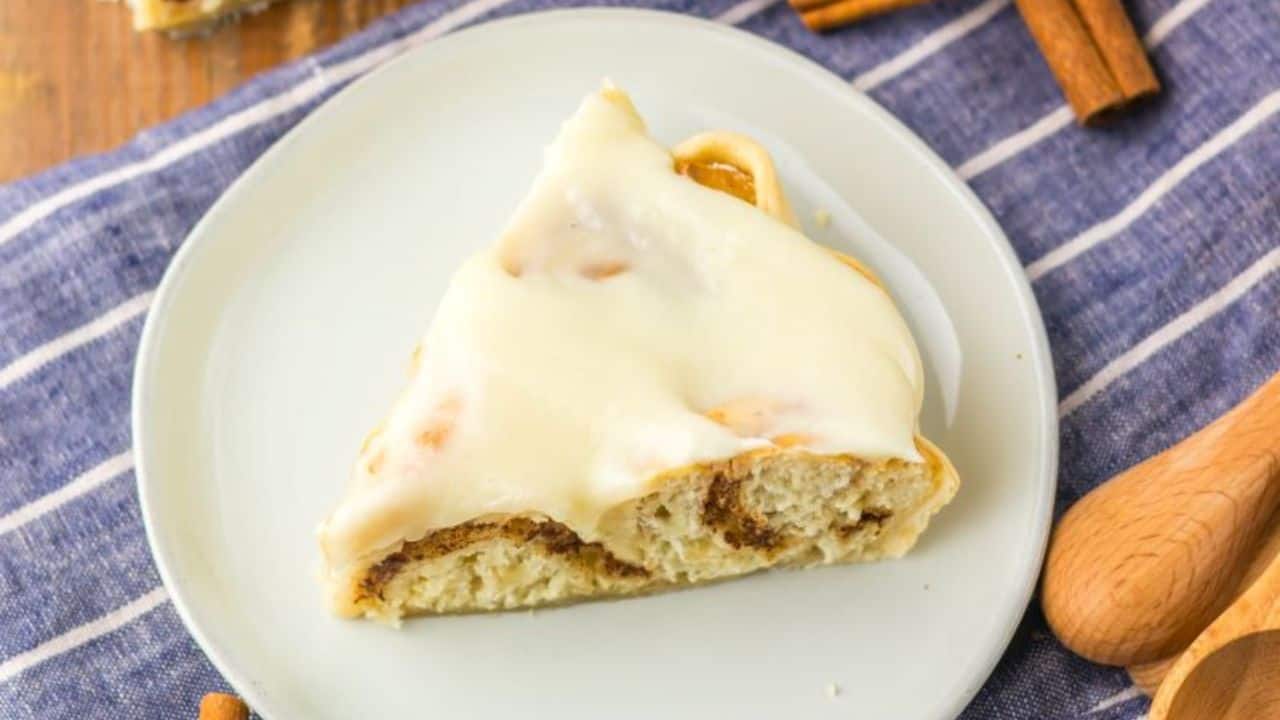 A slice of cinnamon roll pie with icing on a white plate, placed on a striped cloth. Cinnamon sticks are nearby.