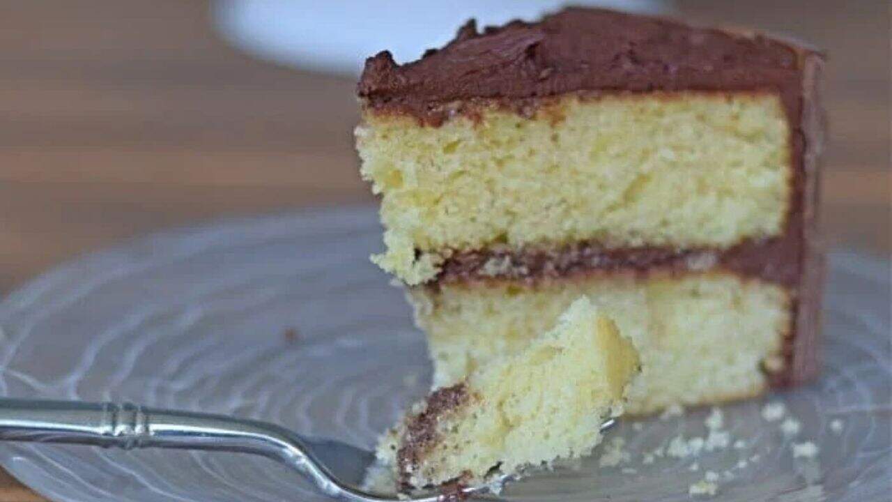 Image shows a slice of yellow cake with chocolate frosting on a plate with the full cake behind it.