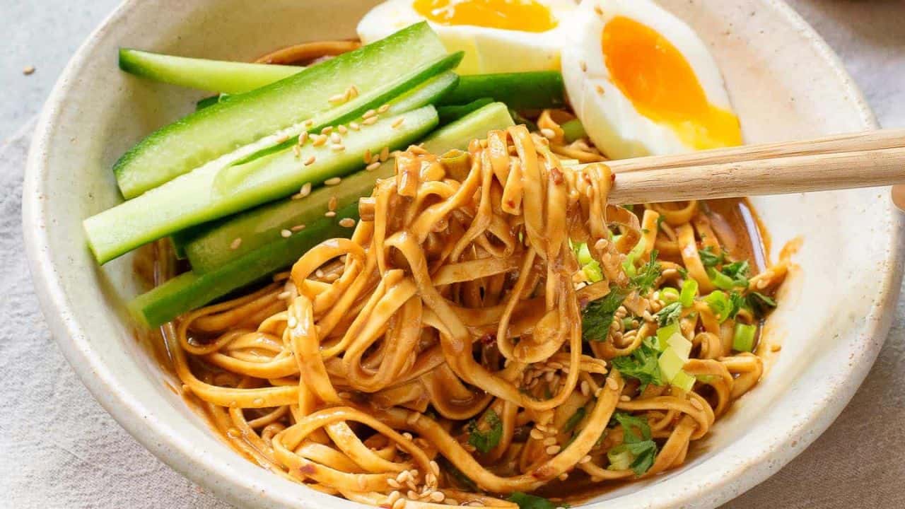 A bowl of noodles topped with cucumber slices, half a boiled egg, and sprinkled with sesame seeds. Chopsticks are lifting some noodles covered in sauce. Fresh cilantro is scattered on top, adding a touch of green.