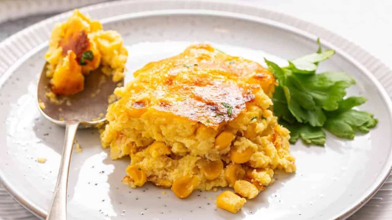 A close up image of a single serving of corn casserole on a plate with a spoon beside it.