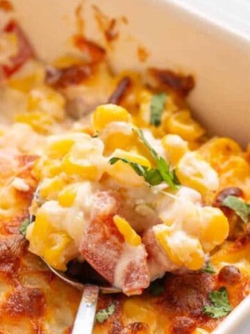 A baked corn casserole with melted cheese and diced red peppers, garnished with chopped herbs, in a white baking dish with a serving spoon.