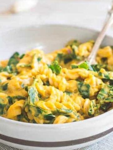 A bowl of pasta with a creamy yellow sauce and spinach, served with a fork on a light, patterned placemat.