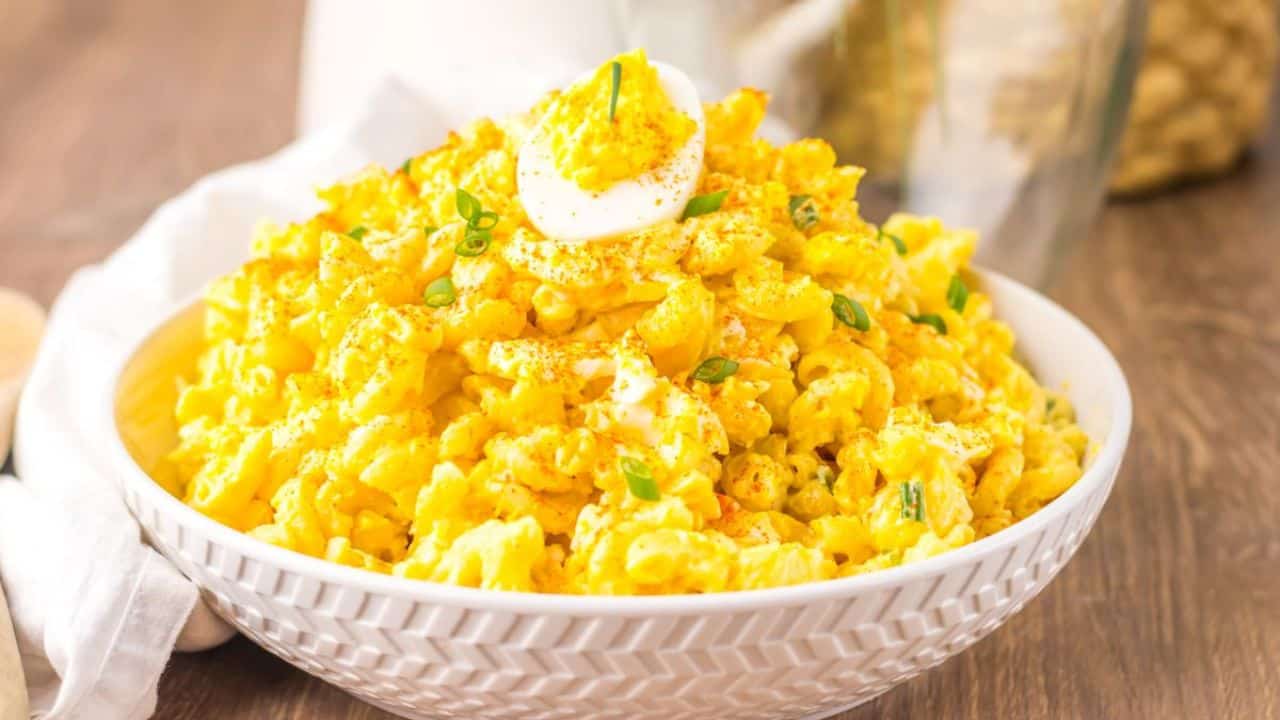 A bowl of creamy macaroni salad topped with sliced boiled eggs, chopped green onions, and a sprinkle of paprika, placed on a wooden surface.