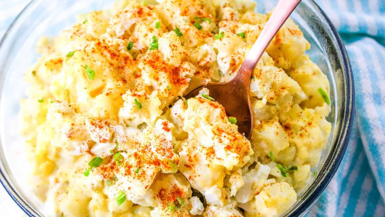 A bowl of potato salad topped with chopped green onions and paprika, with a spoon resting inside.