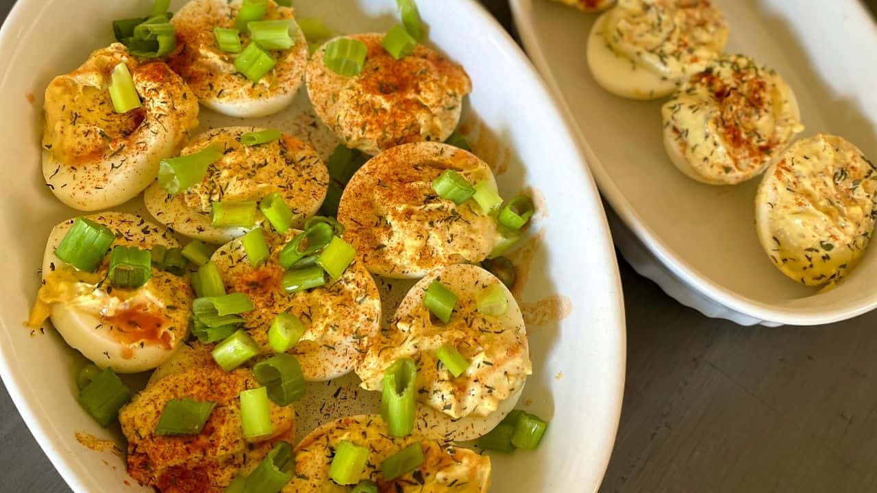 Two white serving dishes with deviled eggs, garnished with chopped green onions, herbs, and a sprinkle of paprika.