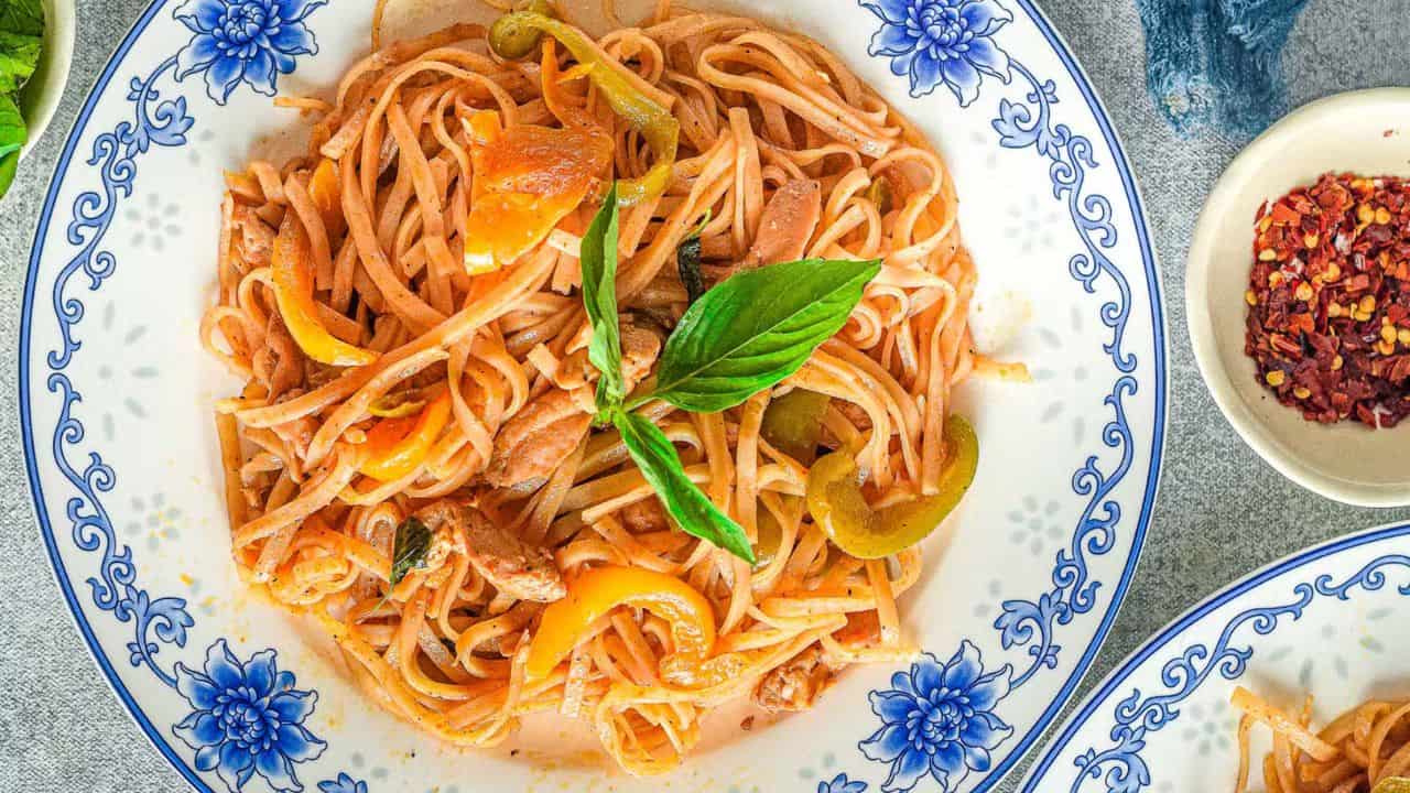 A close-up of a fork holding a portion of pasta with pieces of meat, bell pepper slices, and a basil leaf. The background is blurred, focusing on the vibrant colors and textures of the food.