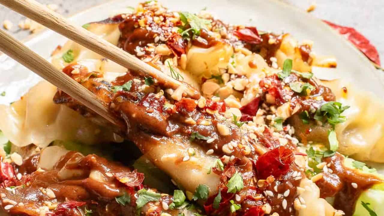 Close-up of dumplings drizzled with a rich sauce, topped with chopped peanuts, chili flakes, and fresh herbs. Chopsticks are picking up one dumpling, showcasing the vibrant toppings and textures.