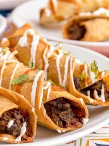 Three beef-filled taquitos topped with sour cream and cilantro are served on a white plate, with more taquitos and a bowl of salsa in the background.