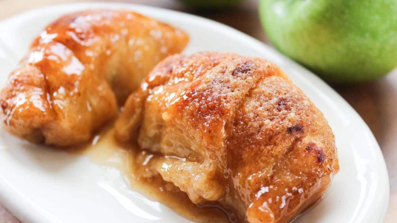 Two baked apple dumplings with a golden-brown crust and syrupy glaze on a white plate, with a green apple in the background.