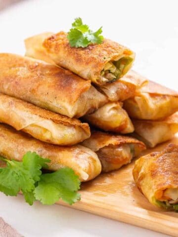 A stack of golden-brown vegetable spring rolls on a wooden board, garnished with cilantro, next to a small bowl of dipping sauce.