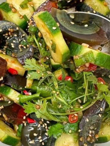 A bowl of century egg salad with sliced cucumber, cilantro, red chili, sesame seeds, and a metal spoon.
