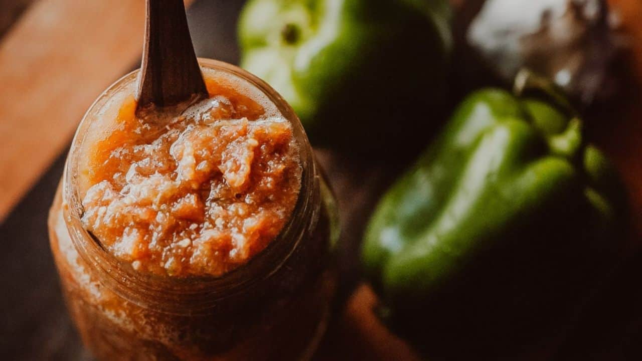 A jar of orange-colored sauce or dip with a spoon, placed beside two green bell peppers and a garlic bulb on a wooden surface.