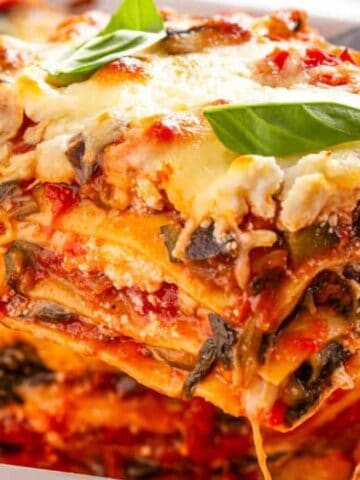 A close-up of a freshly baked vegetable lasagna slice being lifted from a pan, showing layers of pasta, cheese, tomato sauce, and vegetables, topped with basil leaves.