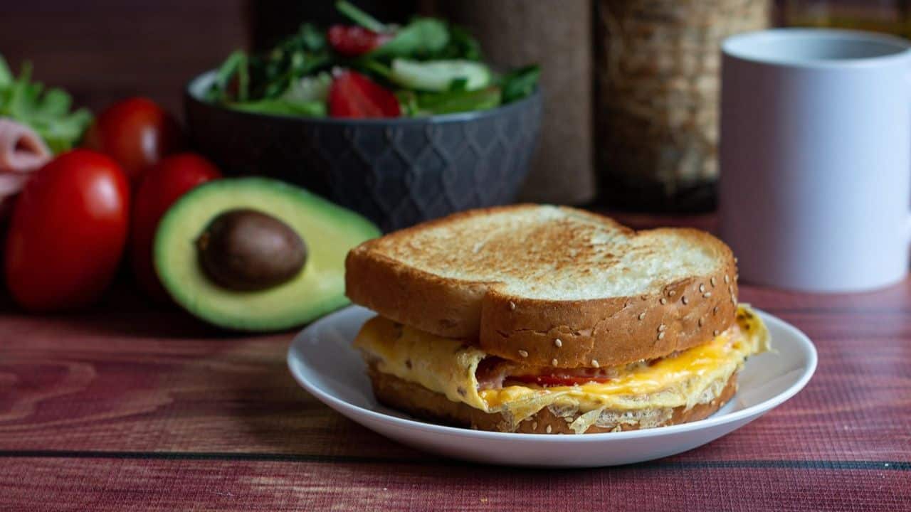 A grilled egg and cheese sandwich on a white plate, with an avocado, tomatoes, a salad bowl, and a white mug in the background on a wooden table.