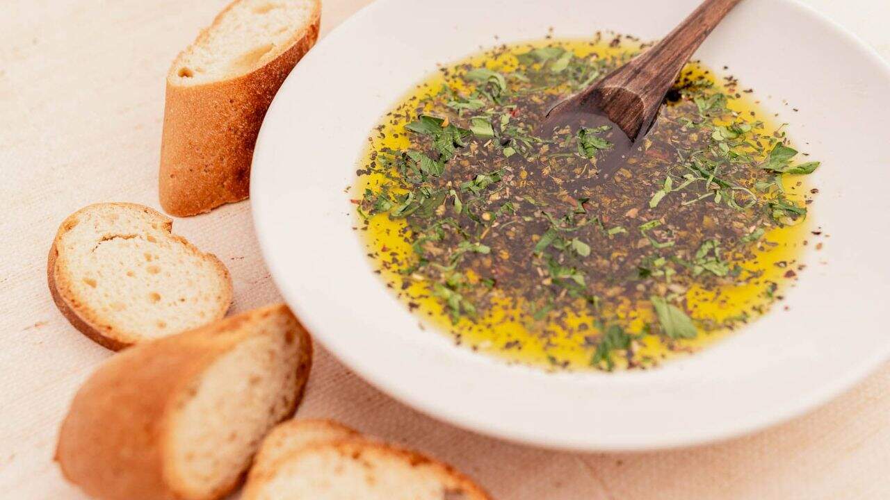 A white bowl of olive oil mixed with herbs and spices, with a wooden spoon, surrounded by sliced baguette on a light surface.