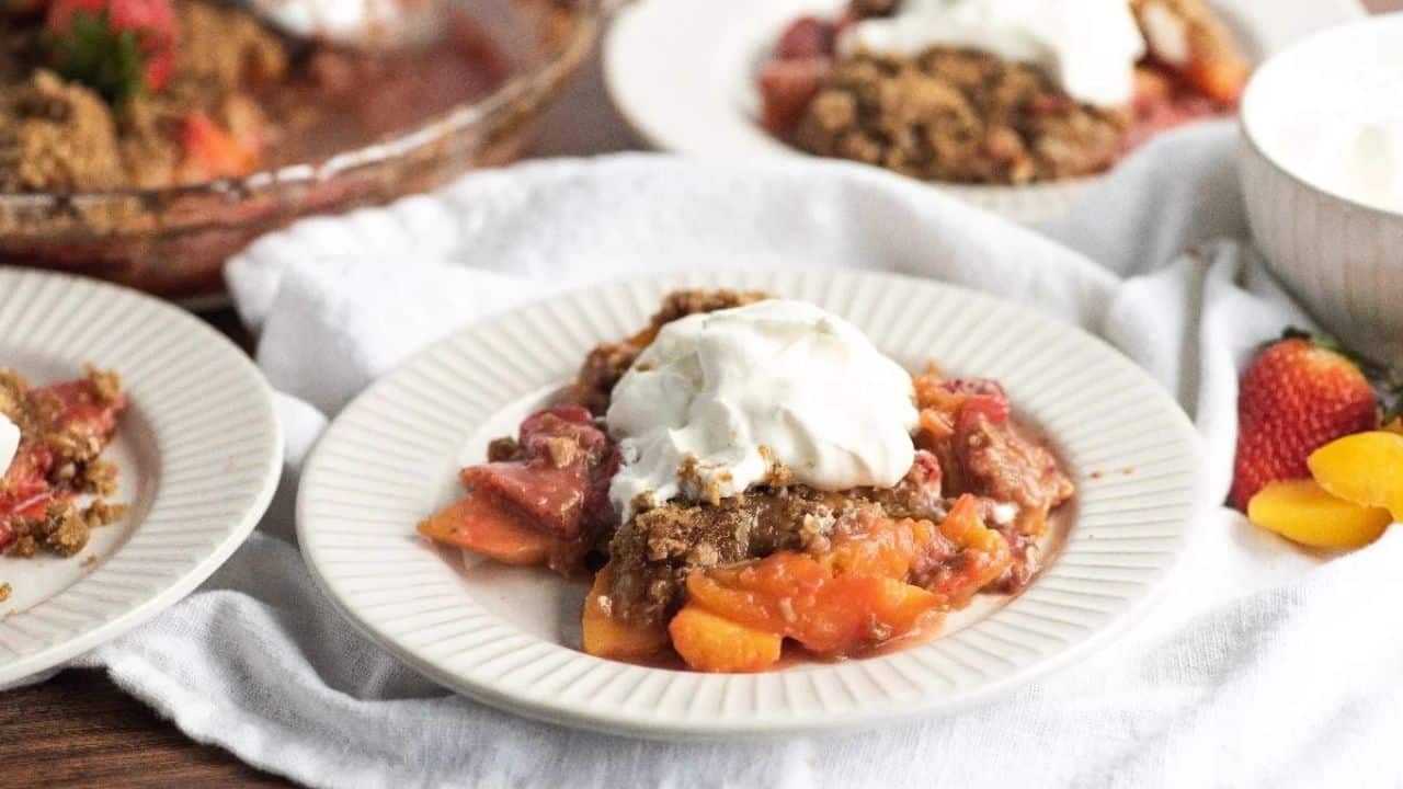 A plate of fruit crisp topped with whipped cream, set on a white dish alongside fresh fruit, with additional servings visible in the background.