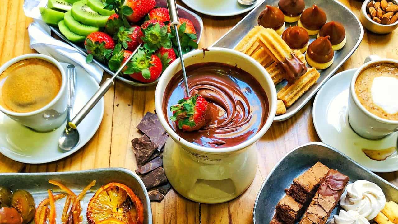 A table set with coffee, brownies, churros, chocolate fondue with strawberries, sliced kiwi, chocolate pieces, and assorted desserts.