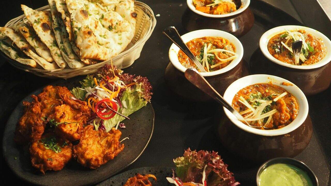 Assorted Indian dishes including naan, vegetable fritters, three bowls of curry, garnished with herbs, and a small bowl of green sauce on a dark surface.