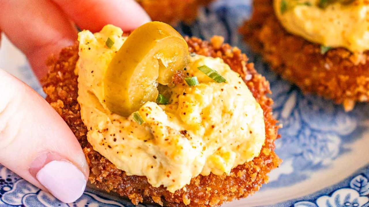A hand holds a fried tomato slice topped with creamy deviled egg mixture, sliced green olive, and chopped chives on a patterned plate.