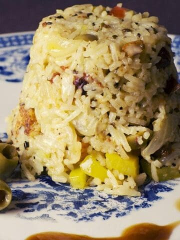 A mound of cooked rice with vegetables and pieces of meat is served on a white and blue plate, accompanied by two green olives and a drizzle of brown sauce.
