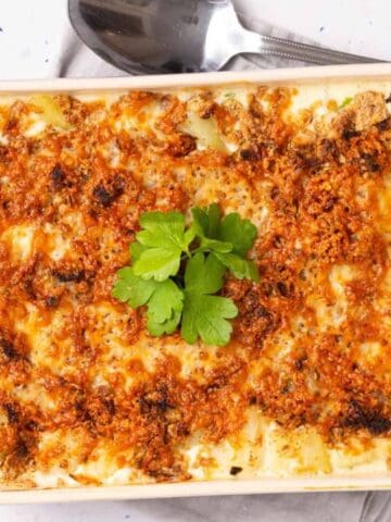 A baked casserole dish topped with golden, crispy breadcrumbs and garnished with fresh parsley, placed beside a spoon and fork on a napkin.