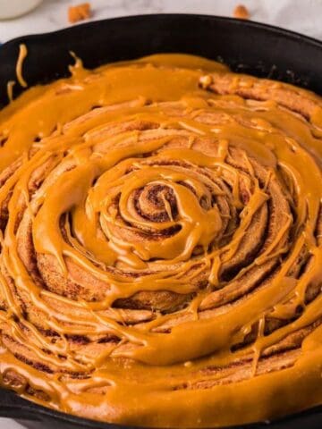 A large cinnamon roll topped with caramel icing in a cast iron skillet, with butterscotch chips and sauce in bowls nearby on a marble surface.