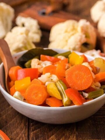 A bowl of mixed pickled vegetables, including carrots, bell peppers, cauliflower, and green beans, sits on a wooden table with jars of pickles and fresh vegetables in the background.