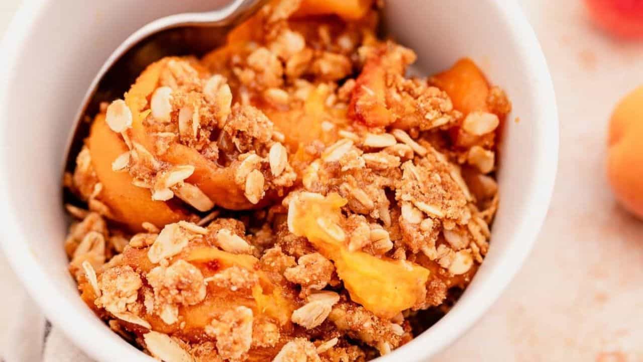 A close-up of peach crisp topped with oats and brown sugar in a white bowl with a spoon.