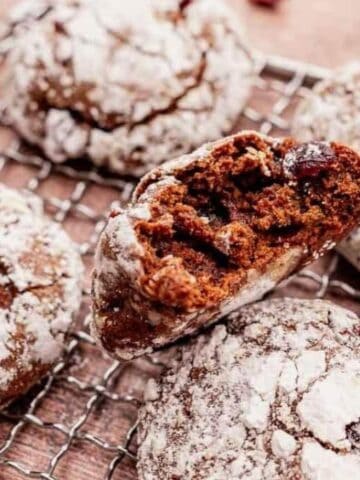 Four chocolate crinkle cookies coated in powdered sugar rest on a wire cooling rack, with one cookie broken in half to reveal a soft, chocolatey interior.