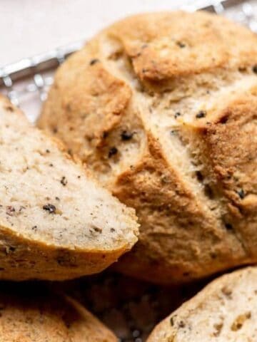 Round loaves of bread with a crisp crust and visible seeds, some whole and one sliced, are resting on a wire cooling rack.