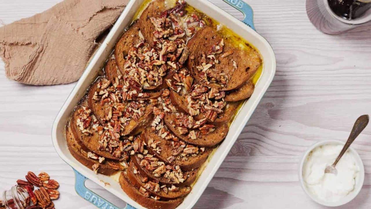 A slice of French toast topped with whipped cream, chopped pecans, and a sprinkle of cinnamon, served on a white plate with syrup pooling around it.