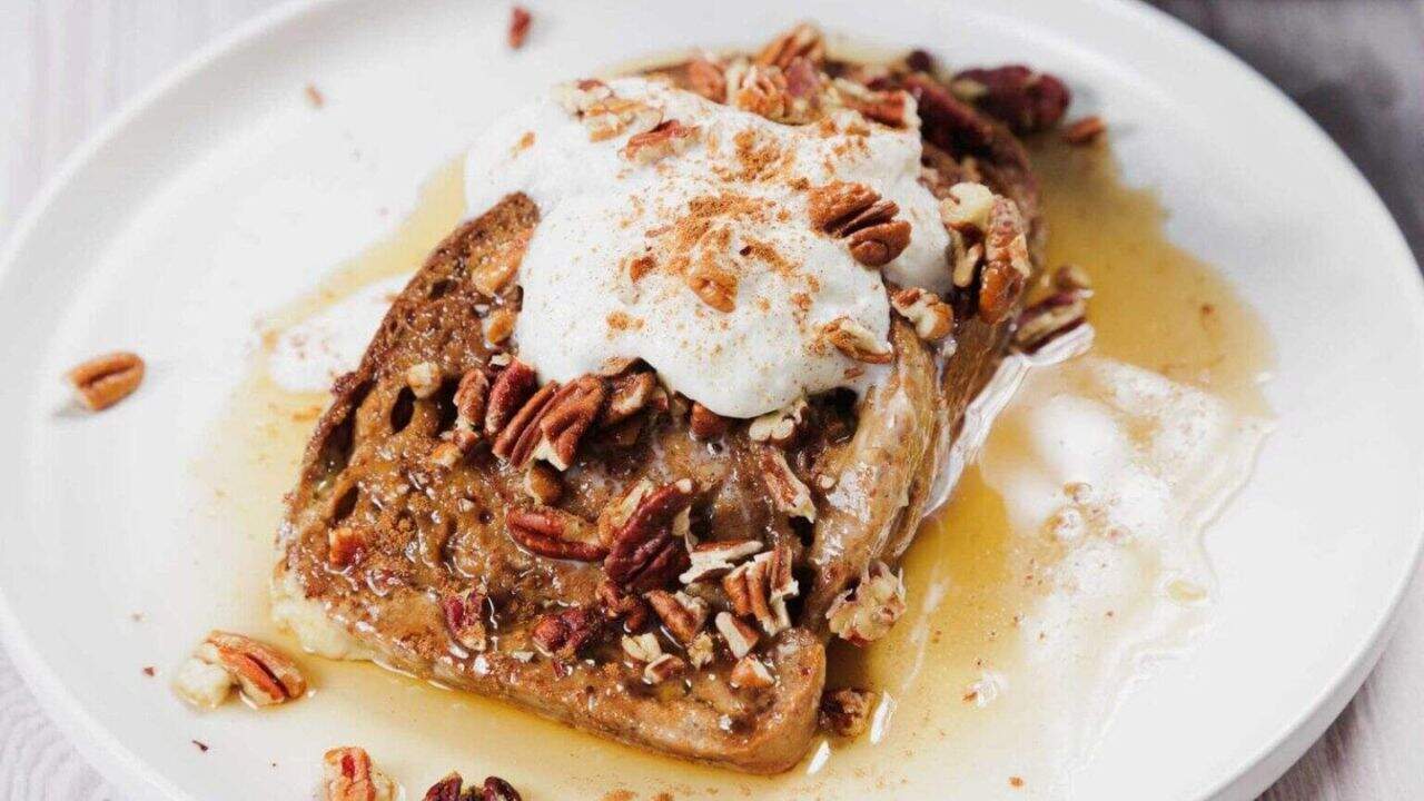 A slice of French toast topped with whipped cream, chopped pecans, and a sprinkle of cinnamon, served on a white plate with syrup pooling around it.