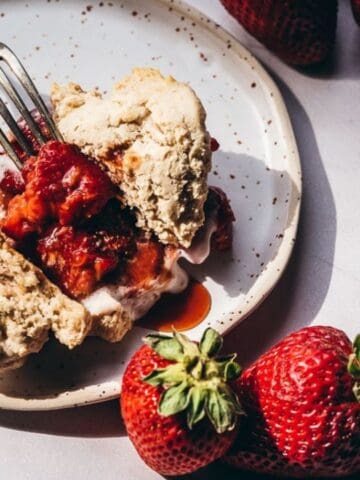A fork rests on a plate with a split biscuit topped with strawberries and sauce, surrounded by fresh strawberries on a white surface.