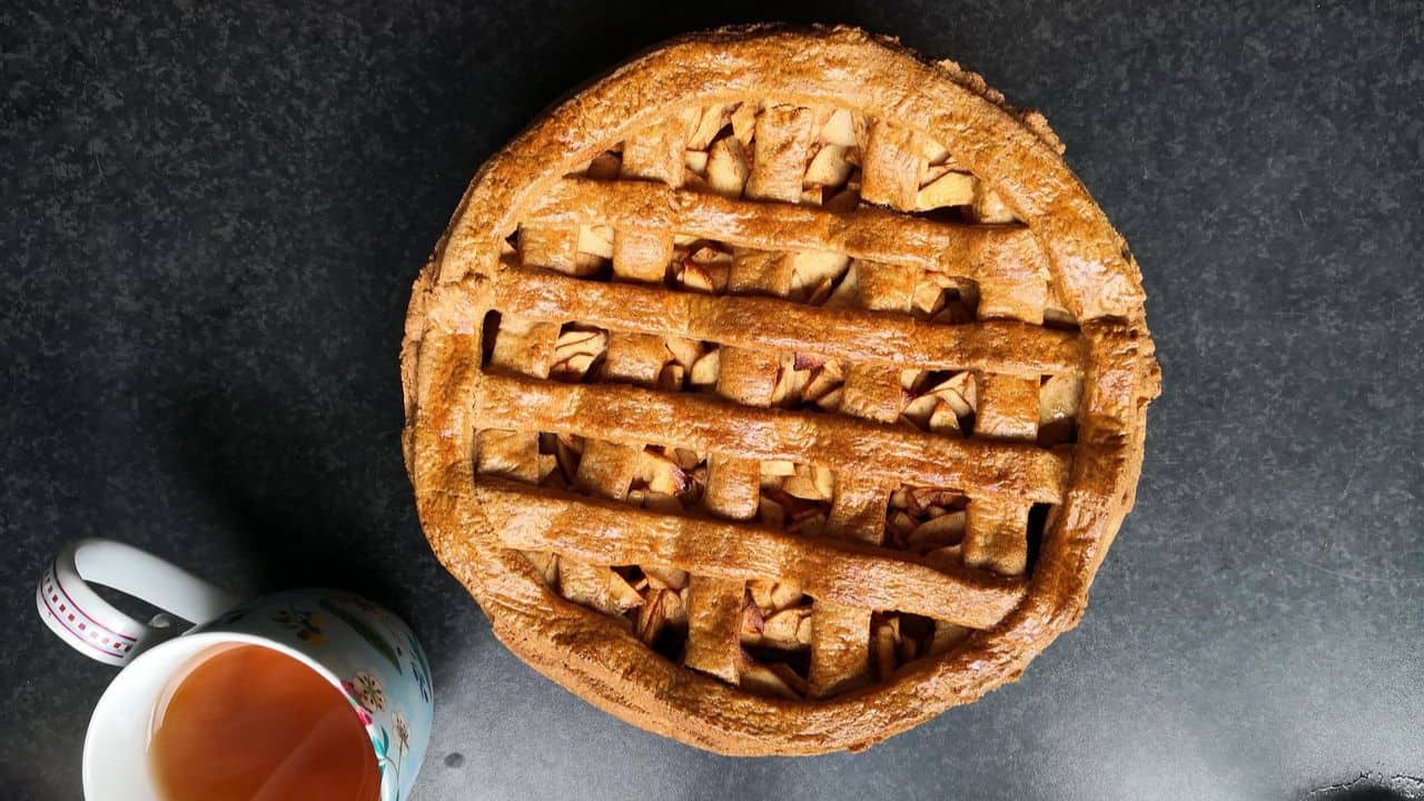 A freshly baked lattice-topped apple pie sits on a dark countertop next to a floral-patterned mug filled with tea.
