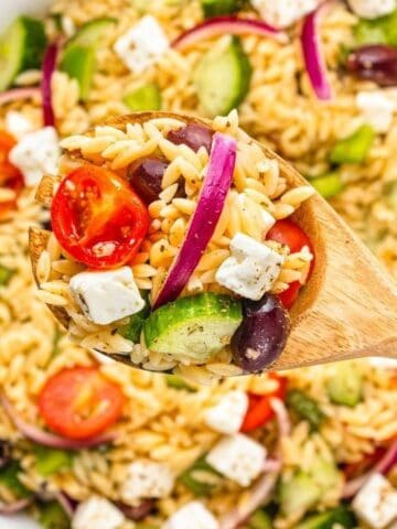 A wooden spoon holds a serving of orzo pasta salad with tomatoes, cucumbers, red onions, olives, and feta cheese above a bowl of the same salad.