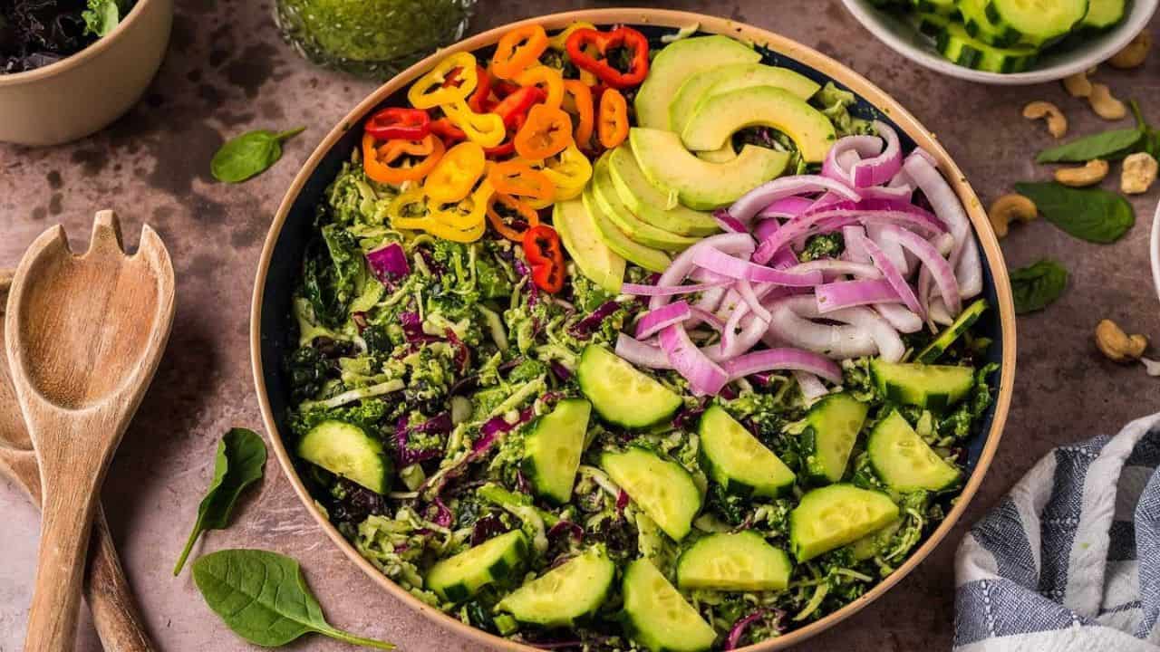 A bowl of salad with sliced cucumbers, red onions, avocado, bell peppers, and greens, arranged in sections on a brown surface with salad utensils nearby.