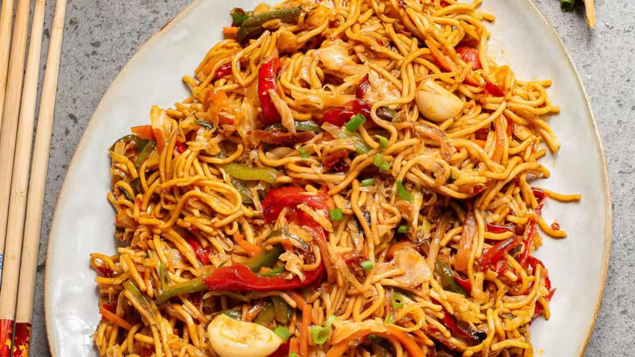 A bowl of stir-fried noodles with colorful vegetables like red and green bell peppers, garnished with spring onions. Chopsticks are lifting a portion of the noodles. A garlic bulb is visible in the background.
