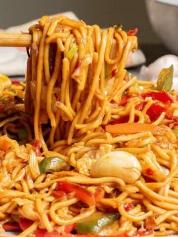 A plate of stir-fried noodles with vegetables is being picked up with chopsticks; a bowl of similar food is in the background.