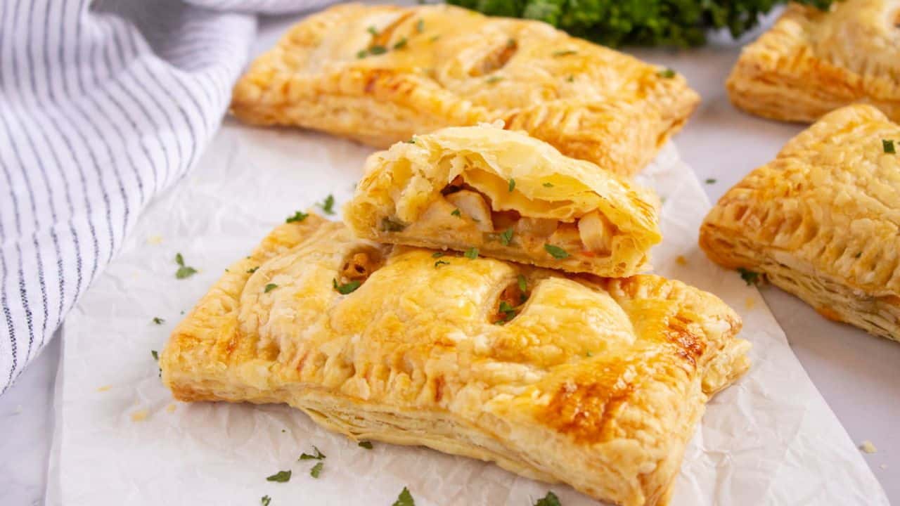 Golden-brown puff pastry pockets filled with a savory mixture of chicken pot pie, placed on white parchment paper, with a striped cloth and green herbs in the background.