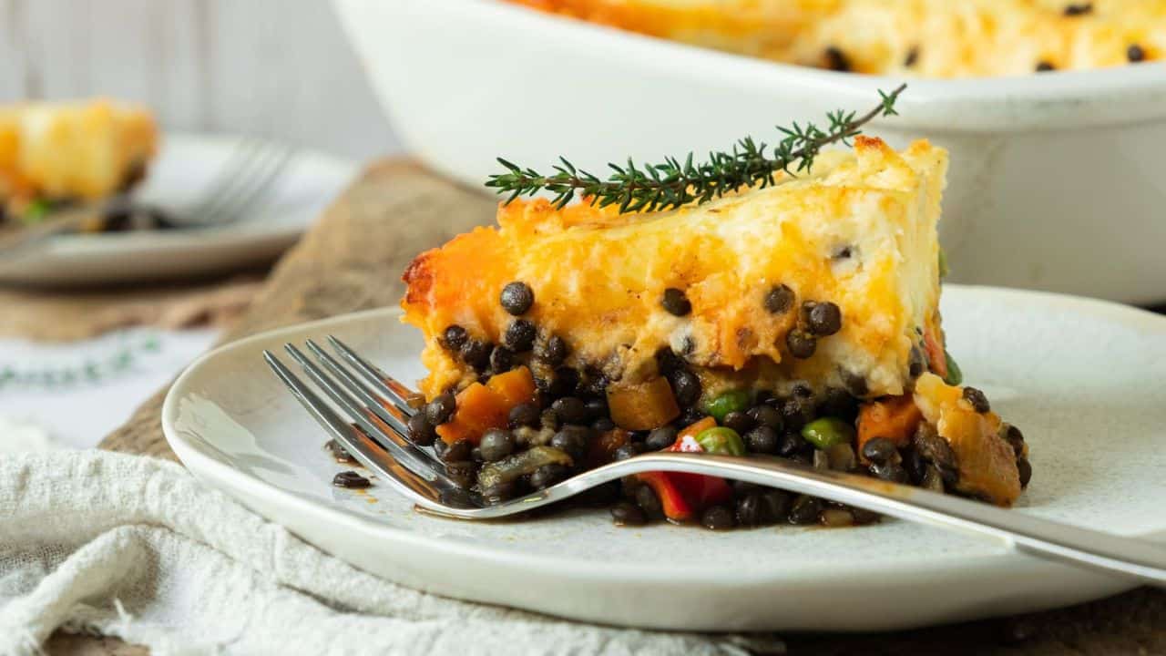 A slice of green lentil shepherd’s pie with mashed potato topping sits elegantly on a plate, garnished with a sprig of thyme.