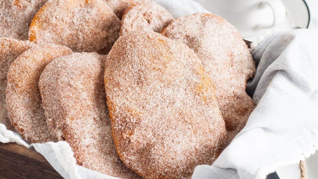 Several pieces of fried dough coated in cinnamon sugar are arranged in a basket lined with a white cloth.