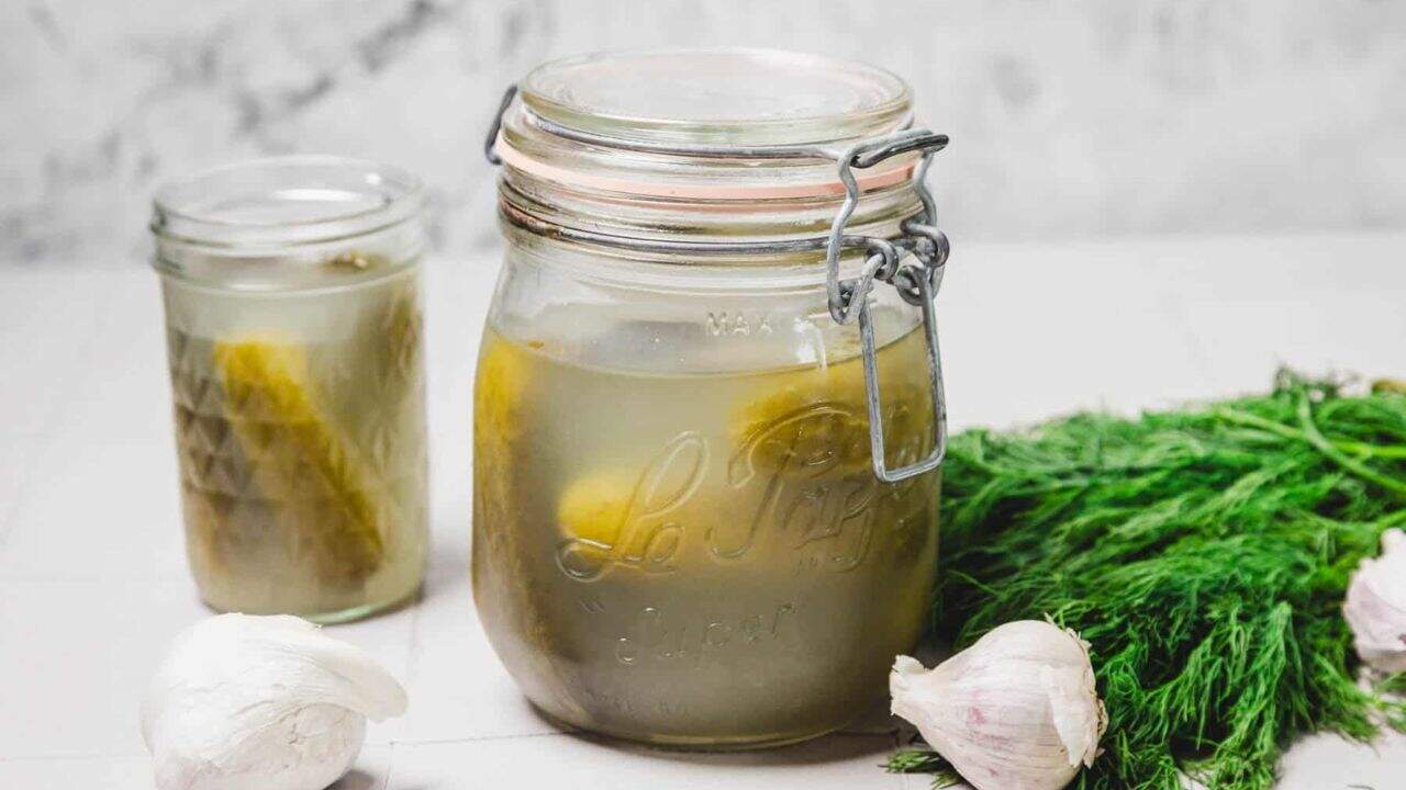 A jar filled with pickles sits on a wooden surface surrounded by ingredients for pickling, including garlic cloves, black peppercorns, and coarse salt.