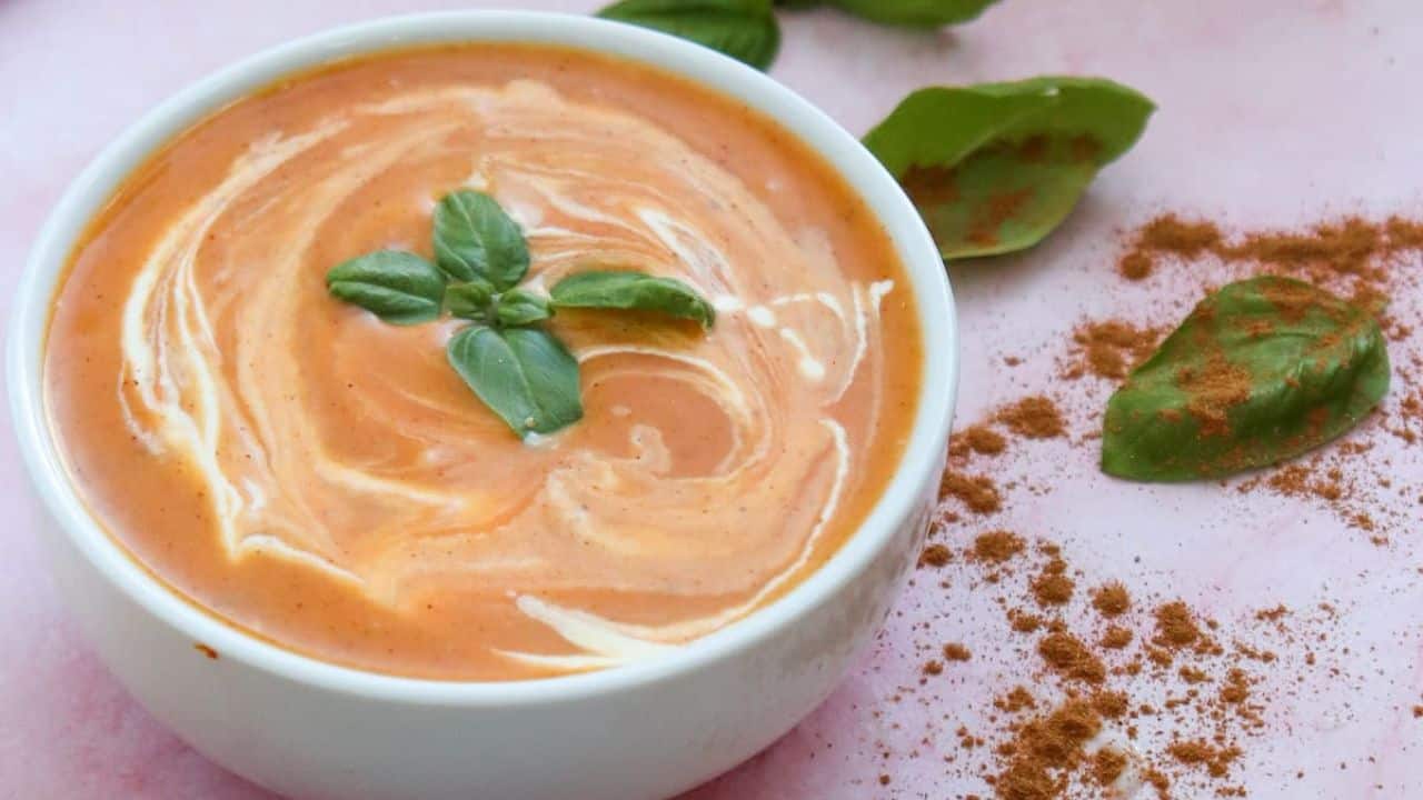 A bowl of pumpkin soup, creamy and garnished with basil leaves, sits invitingly next to a spoon with spices, a whole pumpkin, and a slice of cheese on a light pink surface.