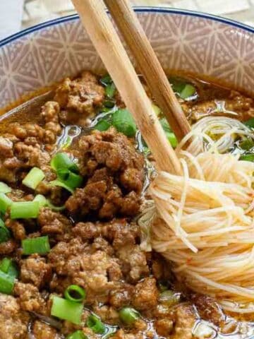Bowl of noodle soup topped with minced meat and chopped green onions, with chopsticks resting on the noodles. Garlic cloves are visible in the background.