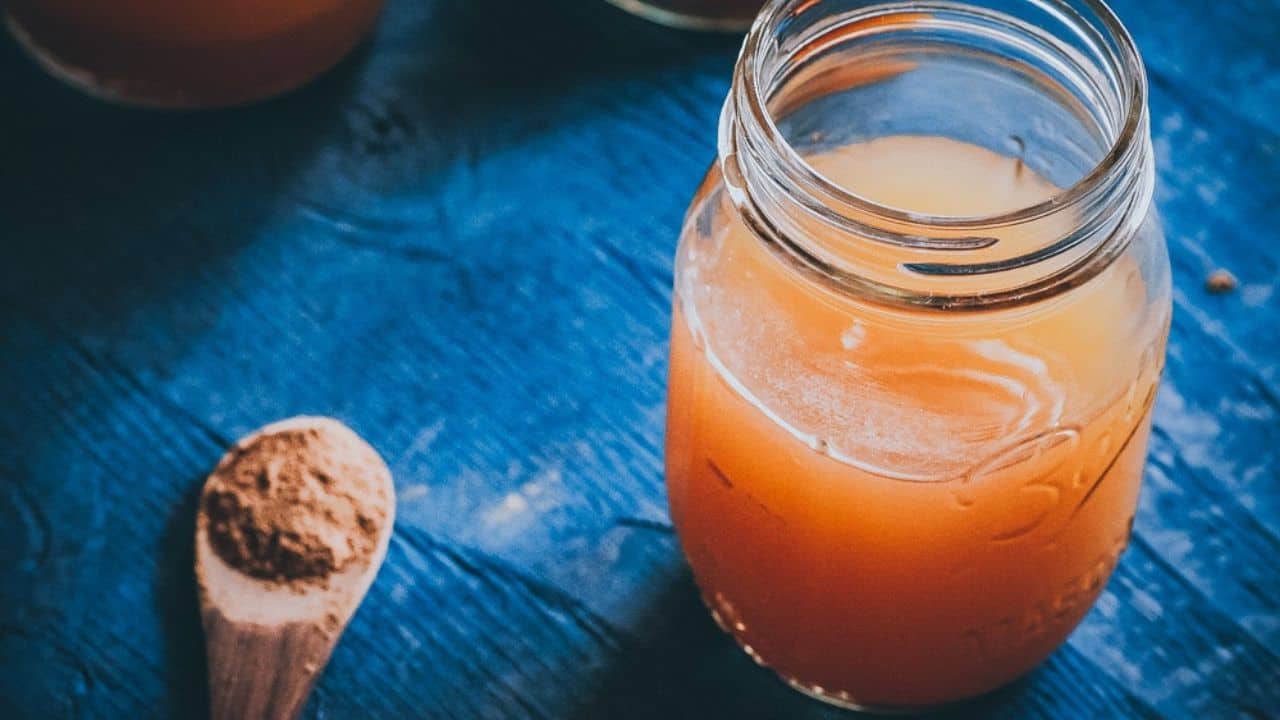 A mason jar filled with orange-brown liquid sits on a blue wooden surface next to a wooden spoon holding ground spices.